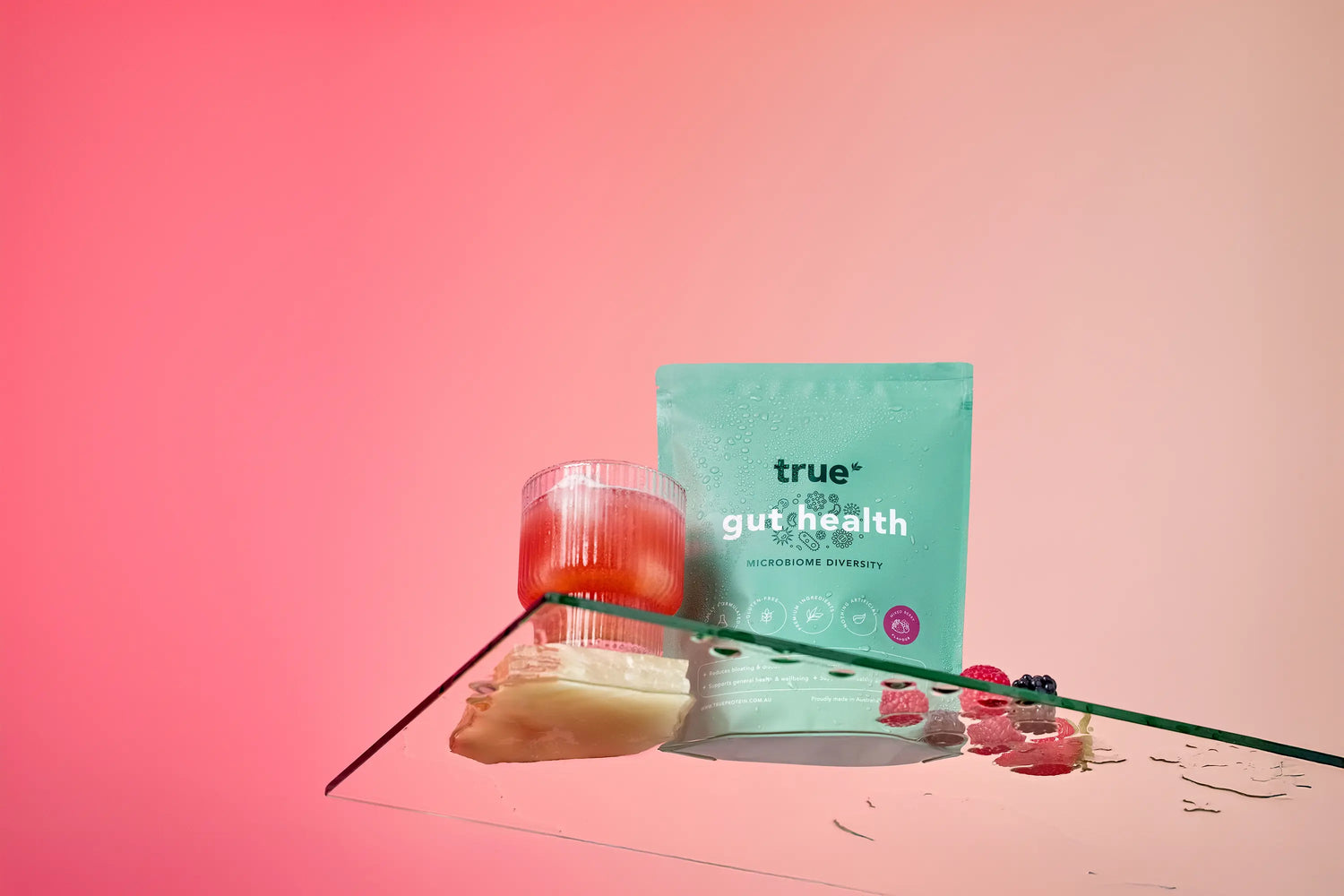 True Gut Health probiotic powder pouch by True displayed with a pink fruit drink in a glass, fresh raspberries and berries and a slice of ginger on a reflective glass surface against a pink gradient background.