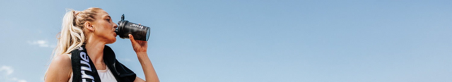 Female athlete drinking from black shaker bottle with towel over her shoulders against a clear blue sky — outdoor fitness/banner image for sports nutrition and workout gear.