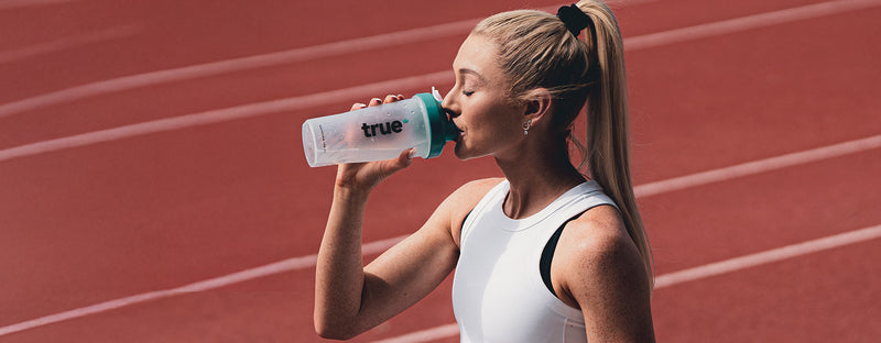 Female athlete drinking from a teal-lidded "true" protein shaker bottle on an outdoor red athletics running track, fitness hydration and sports supplement banner image.