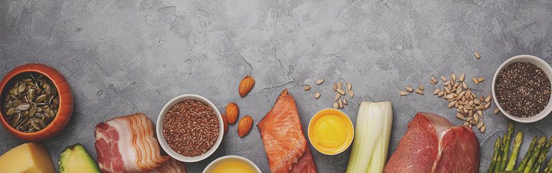 Keto-friendly food assortment on concrete background: salmon fillet, raw beef, bacon, avocado, cheese, eggs, asparagus, celery, almonds, pumpkin seeds, flaxseed and chia seeds.