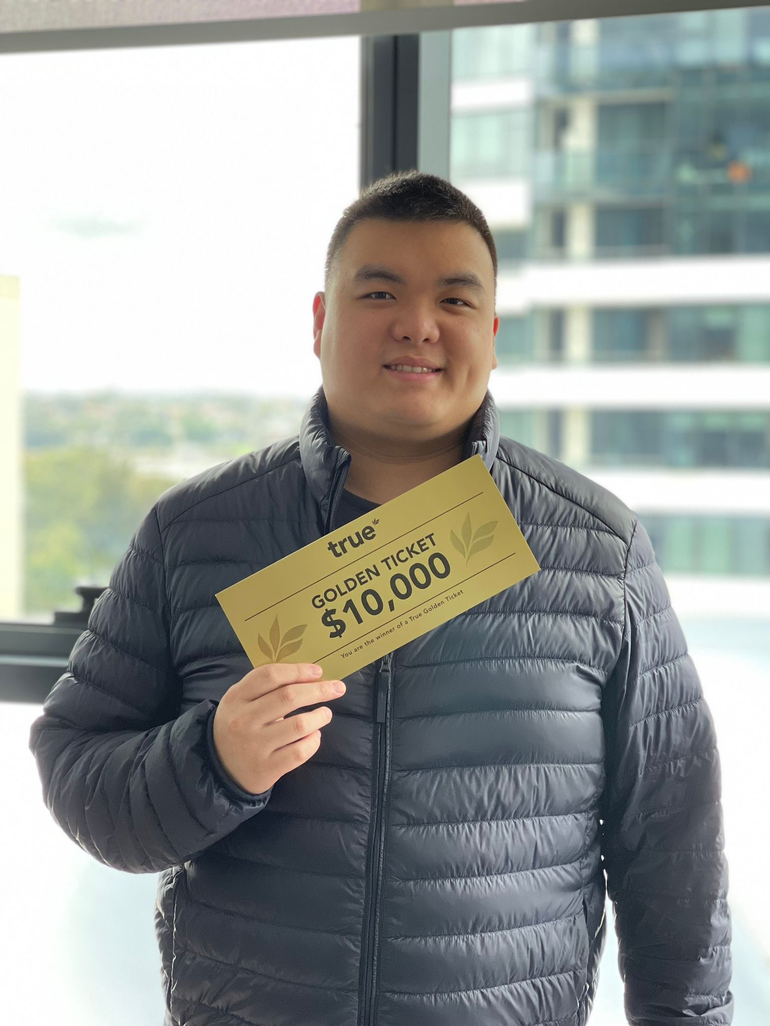 Person in a black puffer jacket holding a gold "true" Golden Ticket that reads "$10,000", standing indoors by a high‑rise window with blurred urban buildings and trees outside.