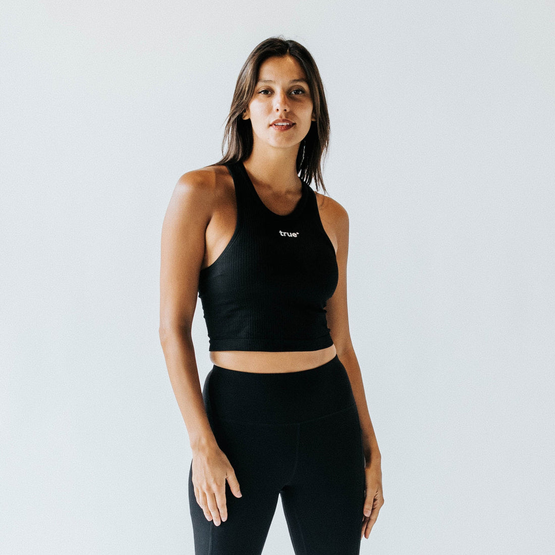 Female model in studio wearing black ribbed racerback crop tank top with small "true" logo and high-waist black leggings — women&