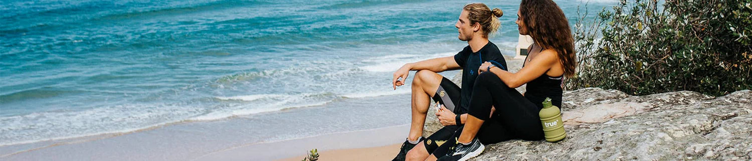 Couple in athletic wear sitting on rocky beach overlooking turquoise ocean waves, sandy shoreline and coastal vegetation with reusable water bottle—outdoor beach workout and recovery scene.