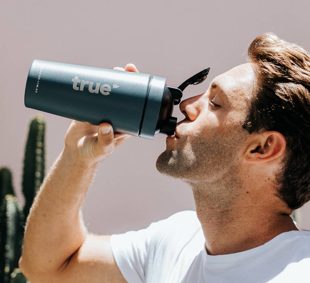 Man drinking from teal "true" insulated water bottle, close-up profile shot against a pale pink wall with cactus in background
