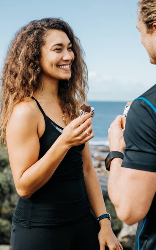 Young woman in black workout clothes smiling and eating a chocolate energy bar while talking with a male runner on an oceanfront coastal trail, healthy snack and outdoor fitness scene.
