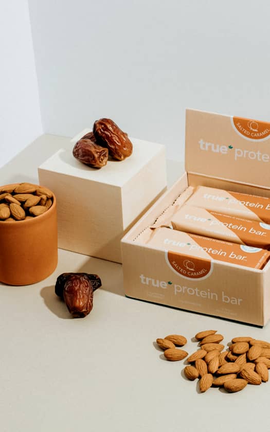Box of True Protein salted caramel protein bars with open packaging, scattered almonds and whole dates on a neutral studio surface — healthy snack ingredients, protein bar product photography.