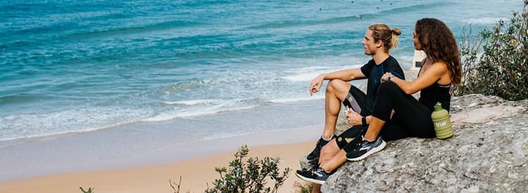 Young athletic couple in workout clothes sitting on a coastal cliff above a sandy beach and blue ocean, with a reusable green water bottle — outdoor fitness and beach view