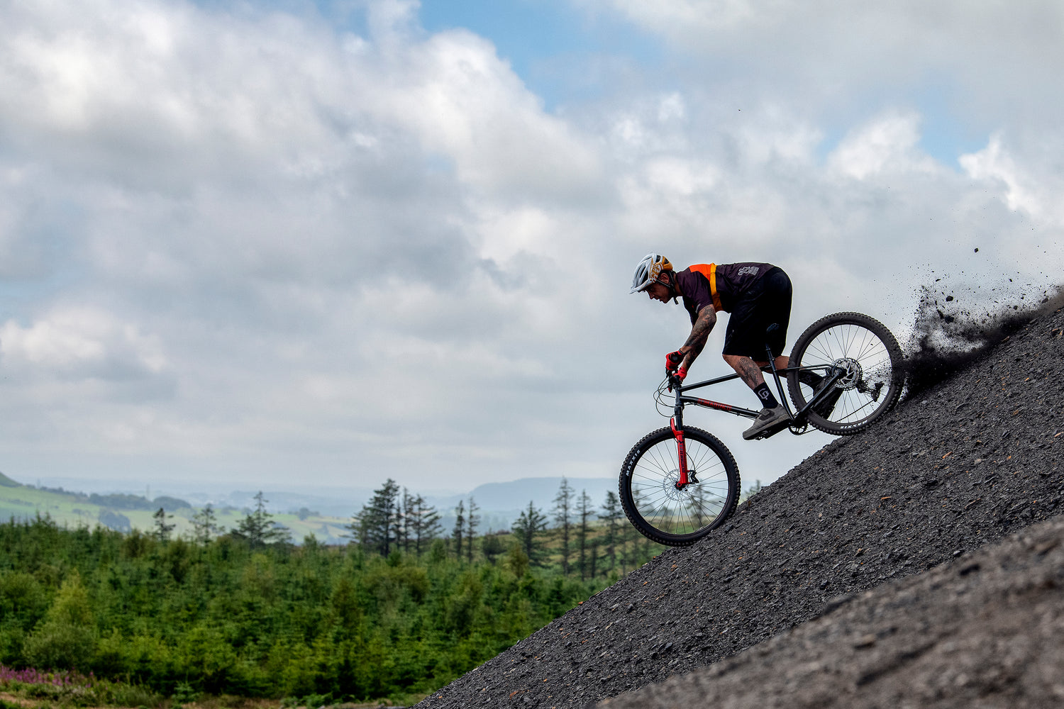 Mountain biker in helmet and gloves riding a red-fronted trail bike downhill on a steep gravel/shale slope, kicking up dust with a green forested upland landscape and cloudy sky in the background — downhill mountain biking action.