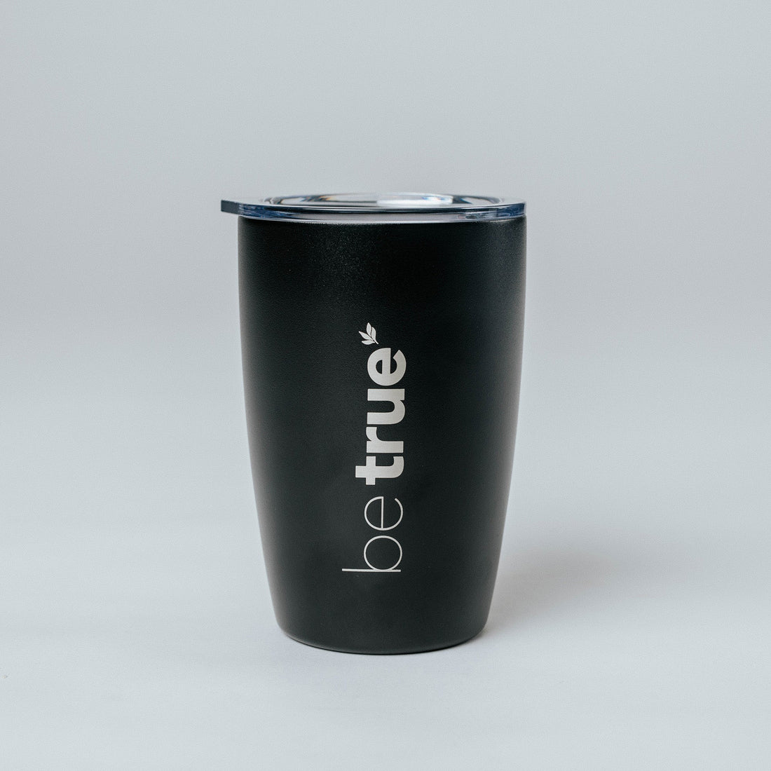 Black insulated stainless-steel travel coffee tumbler with clear lid and vertical "be true" logo, product shot on light gray background — reusable travel mug for coffee and tea.