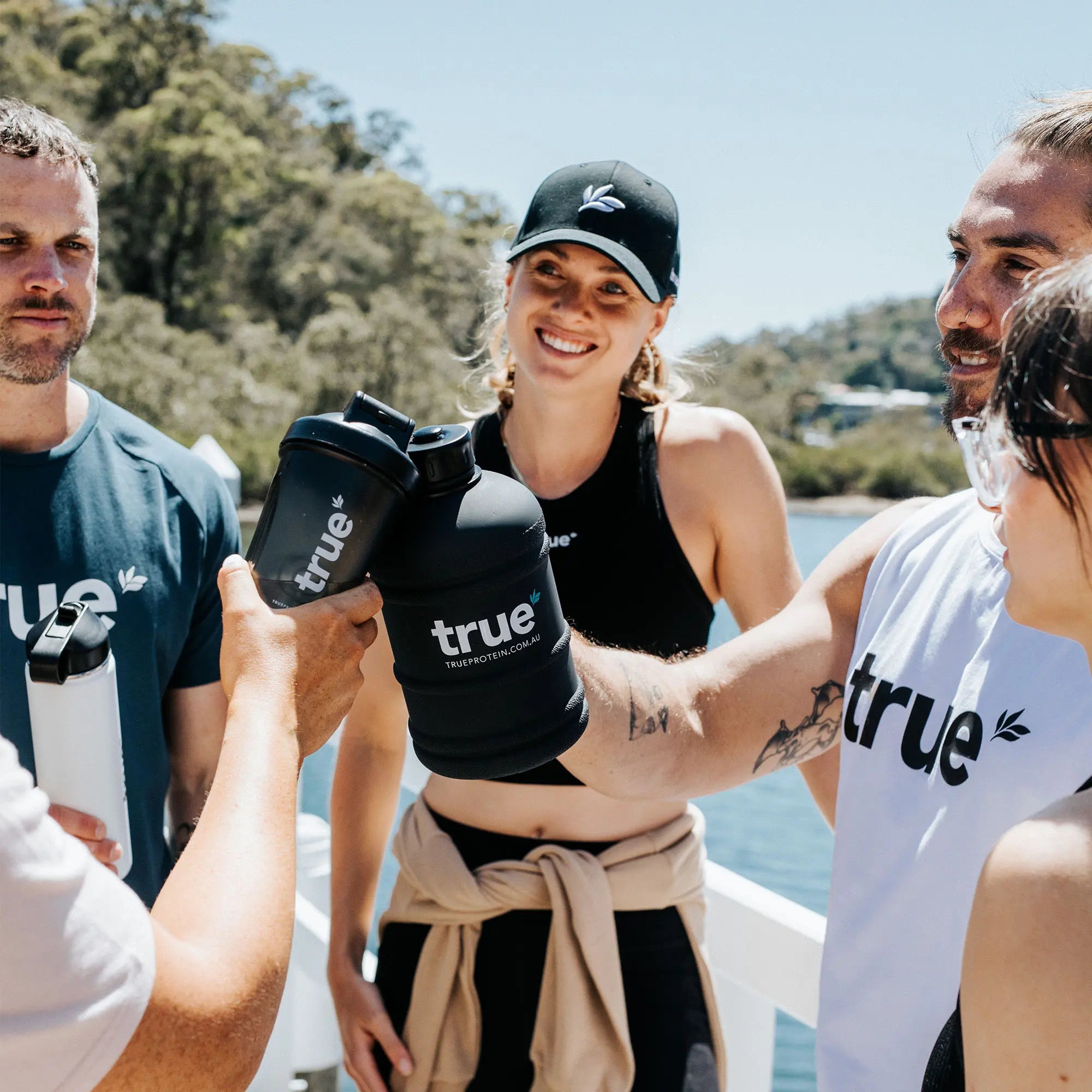 Group of young adults on an Australian waterfront clinking black True Protein shakers and wearing True-branded activewear—outdoor fitness lifestyle, protein shaker bottles and gym apparel.