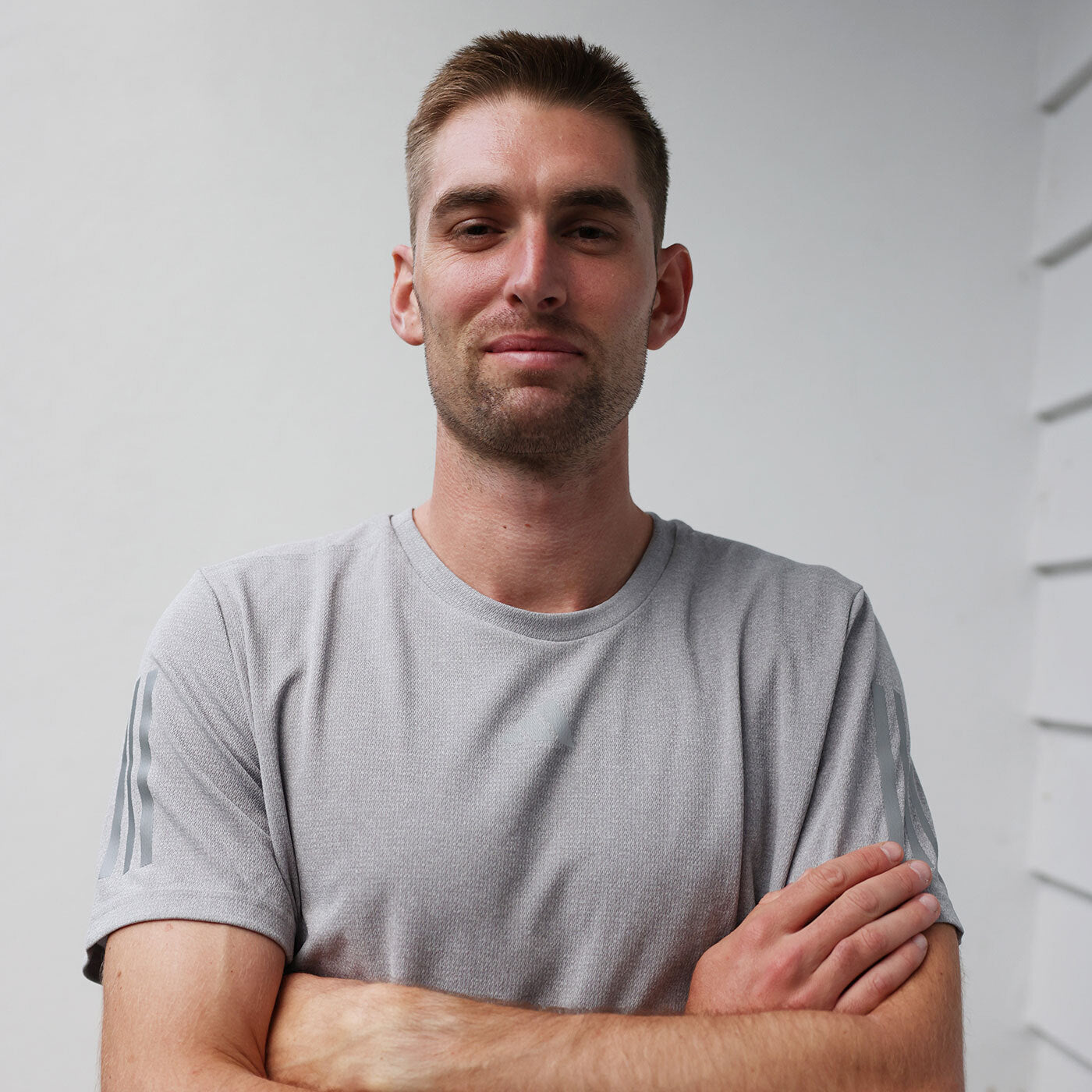 Head-and-shoulders portrait of a young man with short brown hair and stubble wearing a light grey athletic t‑shirt with crossed arms, standing against a white exterior wall in natural daylight — casual male model lifestyle/fitness image.