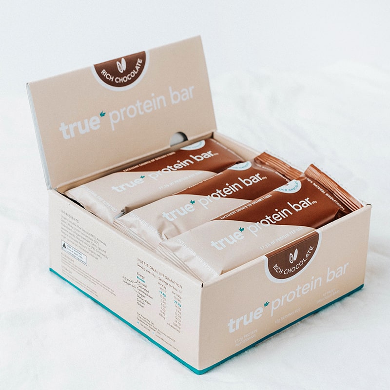 Open retail box of True Protein Bar — Rich Chocolate flavor, individually wrapped protein bars arranged inside on a white background.