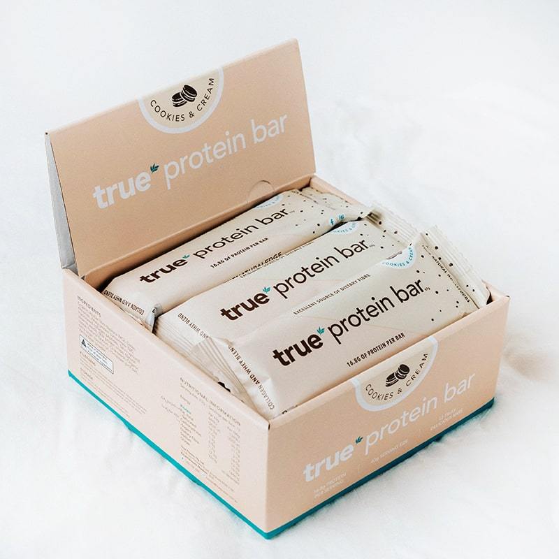 Box of True Protein Bar (Cookies & Cream flavor) with multiple individually wrapped beige bars displayed in an open retail carton on a white surface.