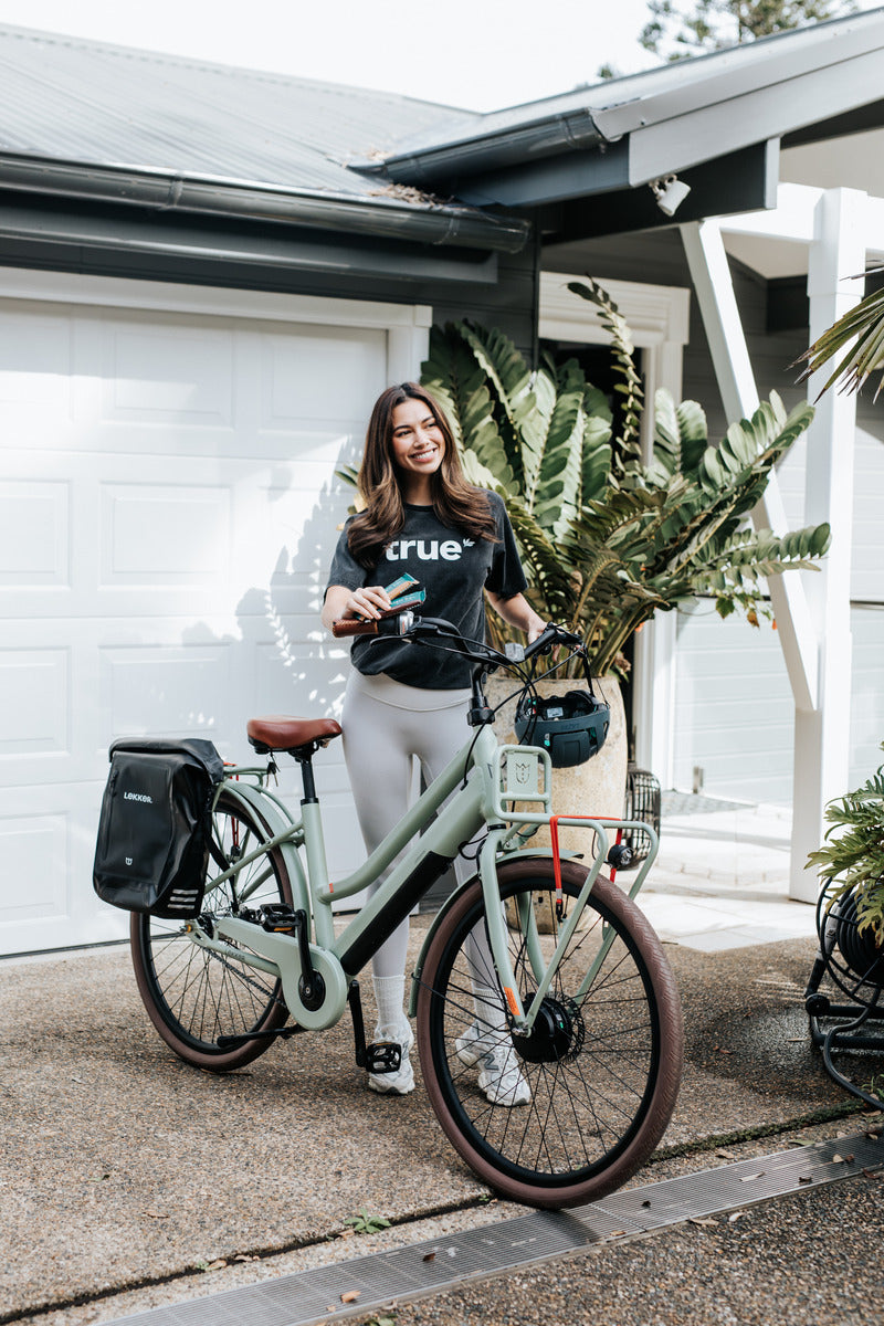 Young woman standing with mint-green commuter electric bike (pannier bag, front rack, helmet) on a suburban driveway outside a modern house — e-bike lifestyle, urban commuter transport, bicycle accessories.