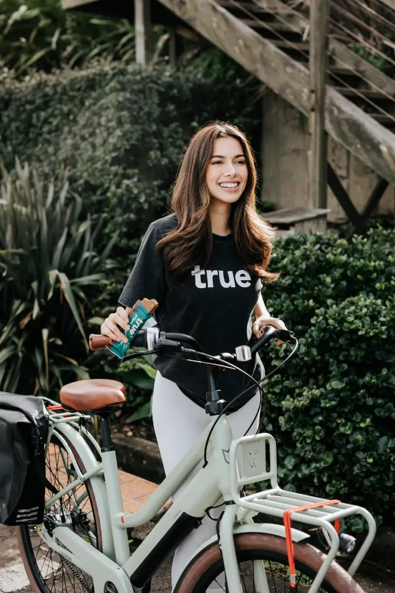 Woman in black "true" t-shirt standing next to a mint green commuter e-bike with front cargo rack and leather saddle, holding a snack bar in a leafy urban garden setting