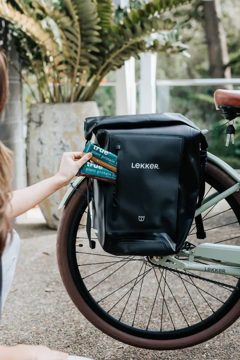 Woman placing green "True plant protein" bars into a black LEKKER waterproof pannier bag on a pale green city e-bike, urban bike commute scene with brown tire and potted plants in background