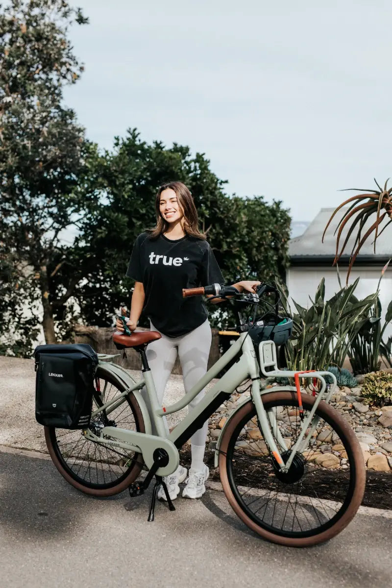Smiling woman in black "true" tee standing with a pale green Lekker electric city bike with rear pannier on a sunny suburban street, ideal for eco-friendly commuting and urban cycling.