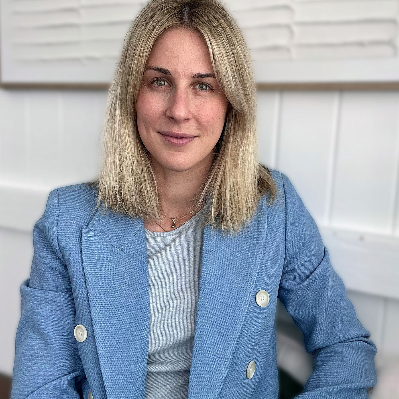 Professional headshot portrait of a blonde woman wearing a light blue double-breasted blazer and grey top, seated against a white paneled interior background — business portrait, corporate headshot, lifestyle branding image.