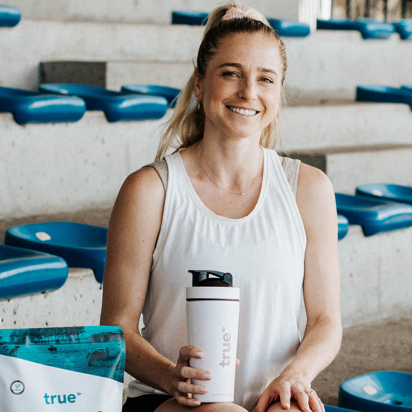 Smiling woman athlete in a white tank top sitting on blue stadium bleachers holding a white "true" branded protein shaker bottle next to a bag of True protein powder — fitness lifestyle product photo.