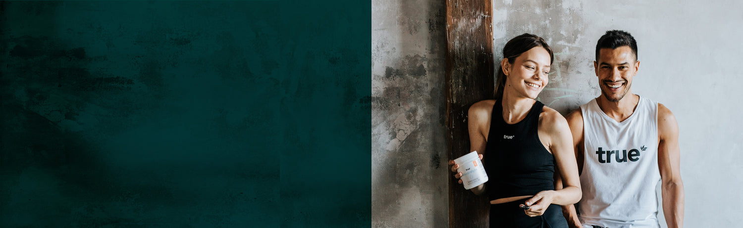 Smiling fit man and woman in workout clothes with "true" logo leaning against a rustic concrete wall in an urban fitness studio, woman holding a True-branded protein supplement tub.