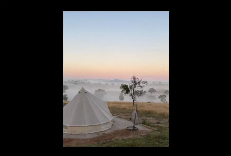 Glamping bell tent on a grassy hill with mist-filled valley and scattered trees at sunrise, peaceful rural campsite dawn scene