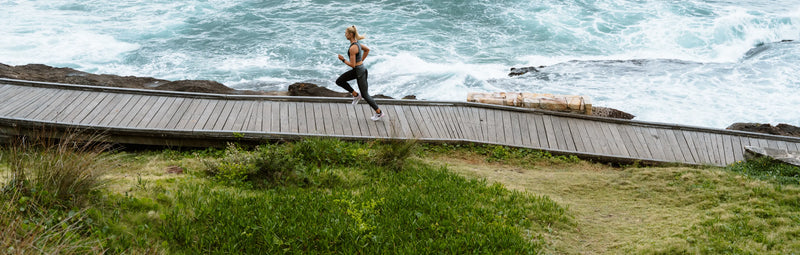 Woman jogging on a wooden coastal boardwalk beside crashing ocean waves — outdoor fitness running along seaside trail, scenic coastal workout setting.