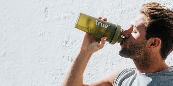 Man drinking from a green "true" reusable water bottle outdoors against a sunlit white wall — lifestyle image for reusable water bottle, sports hydration bottle, fitness accessory