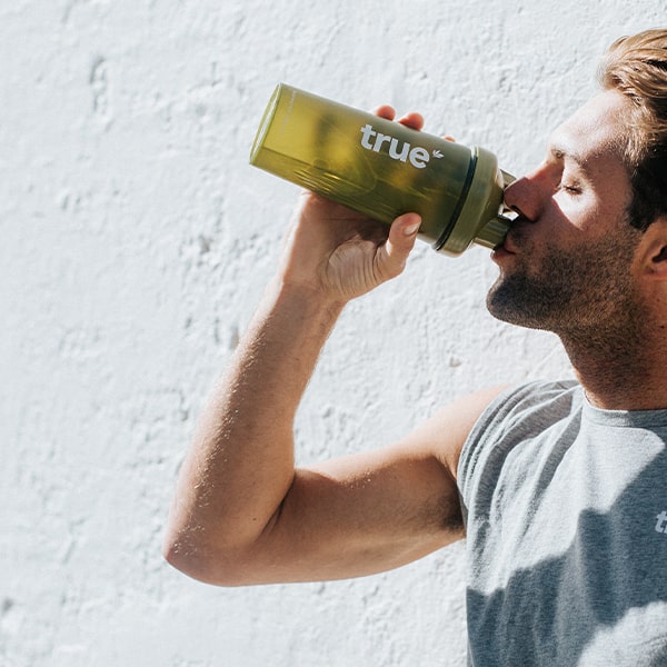 Man drinking from an olive-green "true" branded shaker bottle against a sunlit white wall — reusable protein shaker/gym water bottle for fitness and active lifestyle.