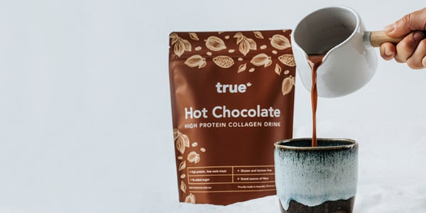 True Hot Chocolate high-protein collagen drink pouch (brown packaging) beside ceramic mug as hot chocolate is poured from a white jug