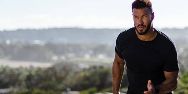 Male runner in a black performance running shirt jogging on a coastal hill trail with blurred seaside landscape in the background — outdoor fitness apparel, men&