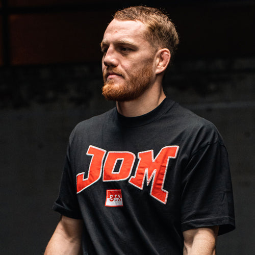 Athletic red‑bearded man wearing a black JDM logo t‑shirt with bold red lettering, portrait on a dark indoor gym/studio background