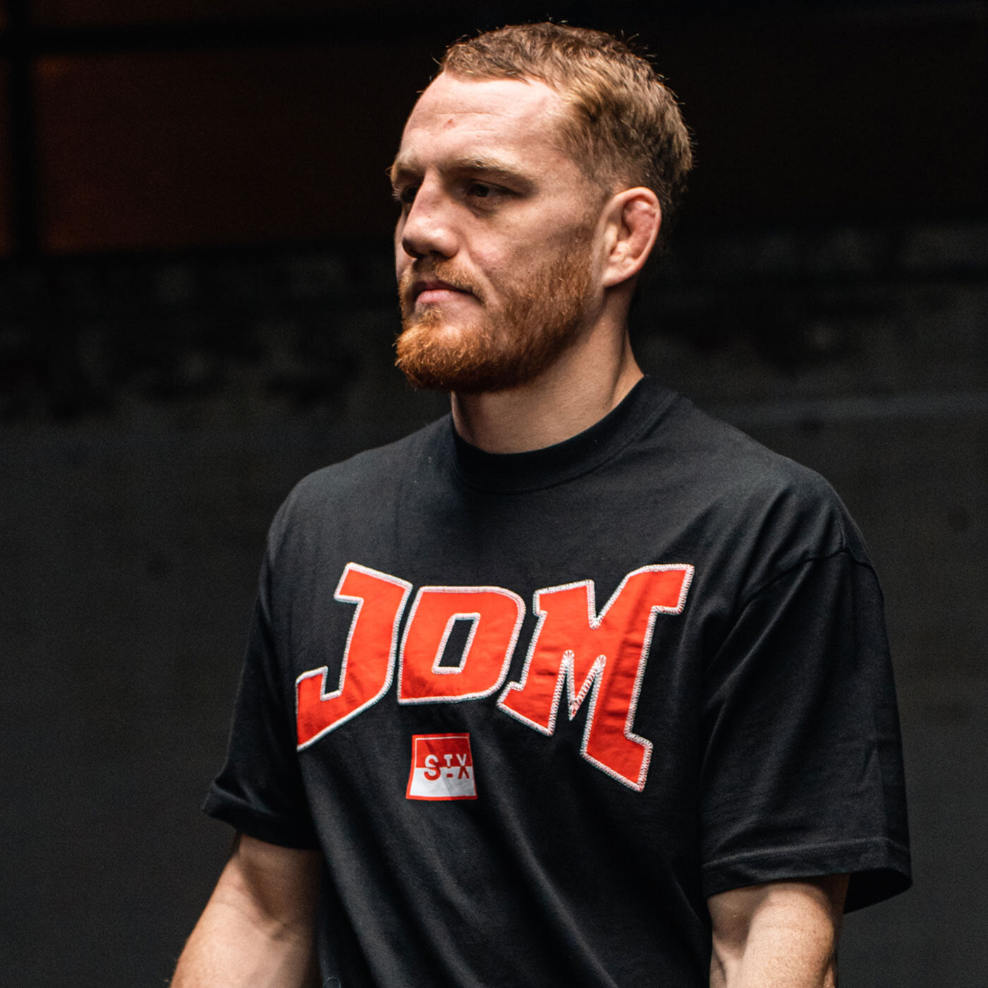 Athletic red‑bearded man wearing a black JDM logo t‑shirt with bold red lettering, portrait on a dark indoor gym/studio background