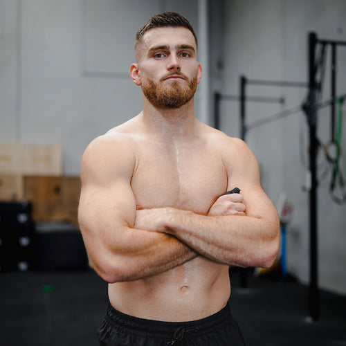 Shirtless bearded male athlete with arms crossed and sweat on chest standing in a CrossFit-style gym with workout rigs and boxes in the blurred background.