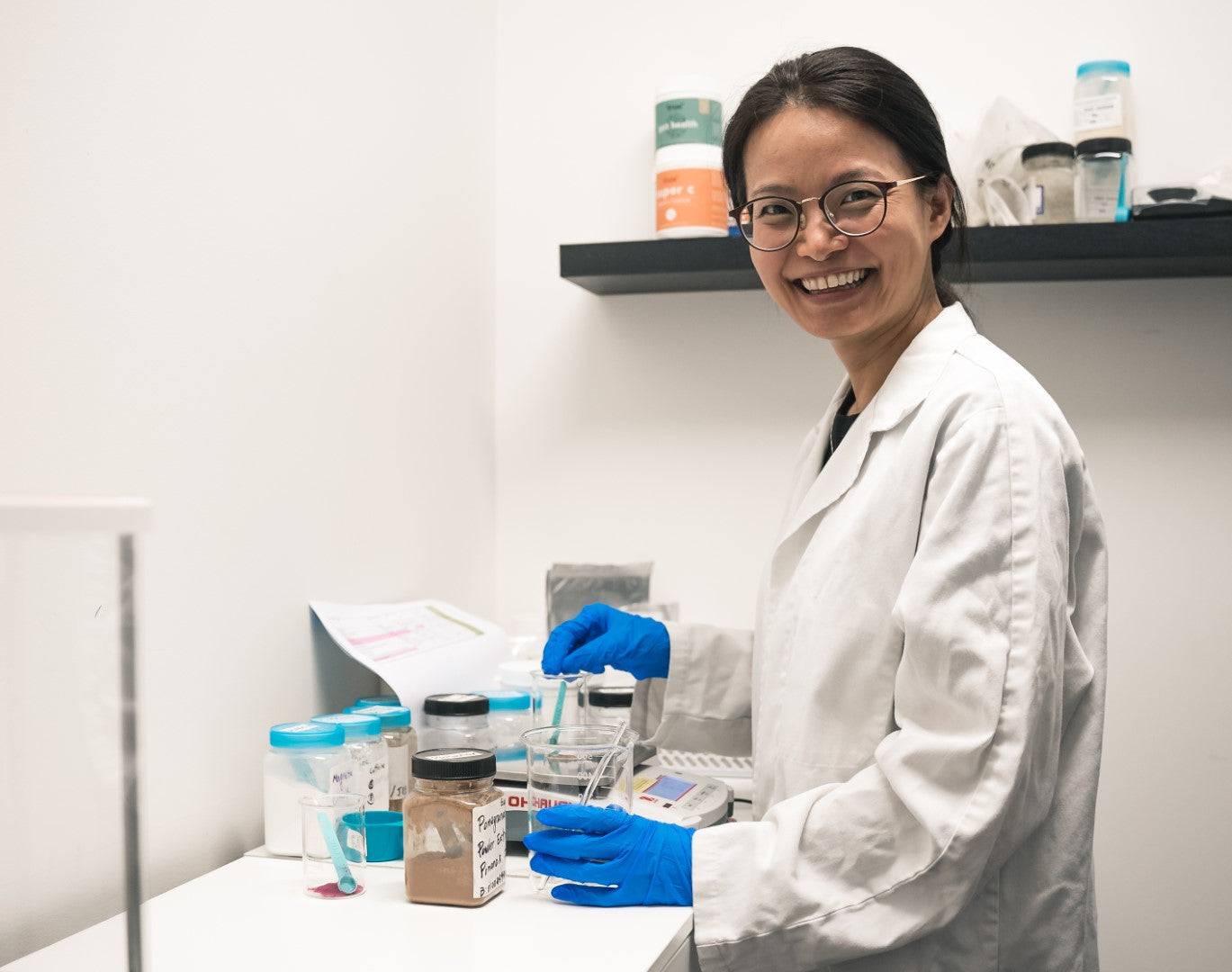 Smiling female lab technician in a white coat and blue gloves measuring and mixing powdered samples in glass beakers on a clinical bench in a modern laboratory, product testing and labeling scene for laboratory services.