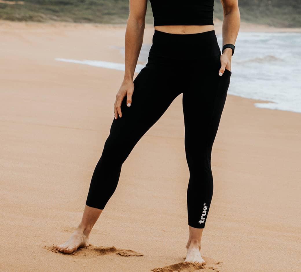Woman wearing black high-waist 7/8 leggings with side pocket and "true" logo, standing barefoot on a sandy beach by the ocean