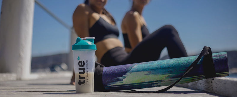True Protein teal‑lid shaker bottle beside a colorful rolled yoga mat on a wooden pier with two women in workout clothes and a blue waterfront sky — outdoor fitness in Australia
