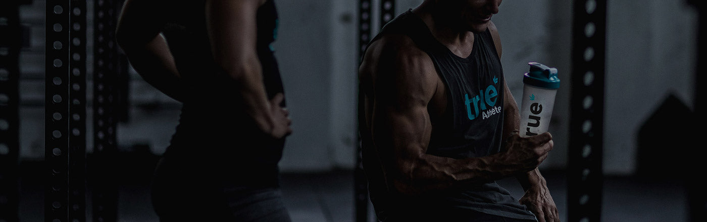 Muscular male athlete in a dim gym holding a branded True Nutrition protein shaker beside a squat rack, strength training and fitness lifestyle banner image