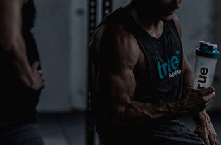 Muscular man in a dim indoor gym holding a True Athlete protein shaker bottle while resting between weightlifting sets