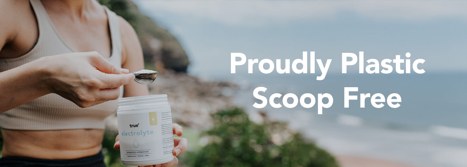 Person at a coastal overlook scooping True electrolyte powder from a scoop-free jar with a spoon; large text overlay reads "Proudly Plastic Scoop Free" over a blurred beach and ocean background