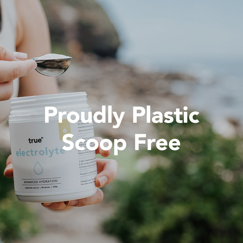 Hand holding a True electrolyte powder jar and metal spoon at a rocky seaside — scoop-free, plastic-free eco-friendly hydration powder packaging.