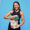 Young woman in a black tank top smiling and holding a stacked bundle of meal kit/food delivery boxes against a bright blue studio background — healthy meal kits, convenient organic meal delivery.