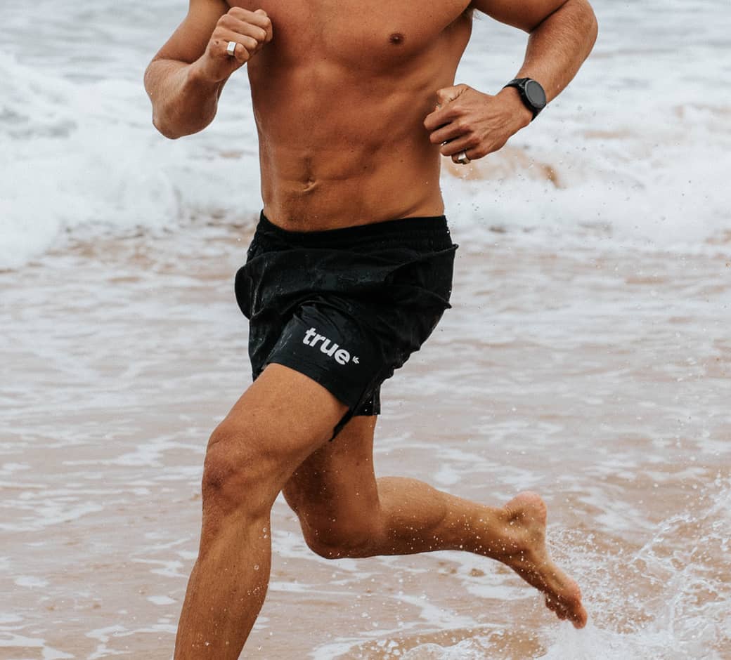 Man wearing black "true" logo men's swim shorts running barefoot through shallow ocean waves on a sandy beach, athletic torso and legs splashing water.