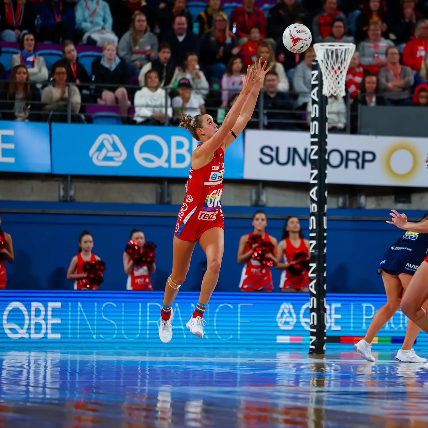 Netball player in red uniform leaping to intercept a shot near the post on an indoor court with QBE and Suncorp advertising boards and a crowd of spectators — Australian professional netball match.
