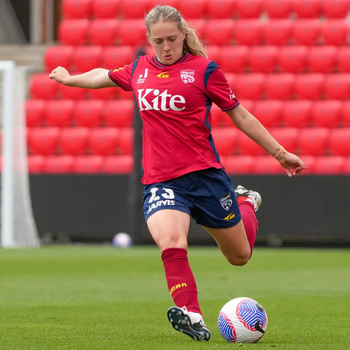 Woman's soccer player in red "Kite" jersey and navy shorts kicking a match ball on a green pitch in front of empty red stadium seats — women's football action shot.