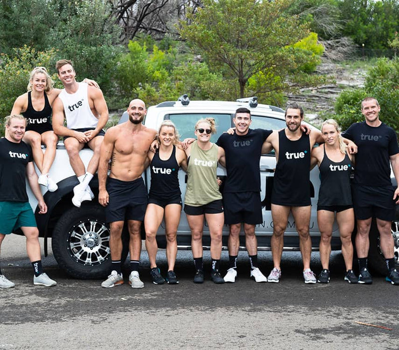 Group of fit mixed-gender trainers in "true" branded workout gear posing in front of a white 4x4 truck on a roadside with coastal scrub and trees — outdoor fitness team, bootcamp trainers, group workout photo.