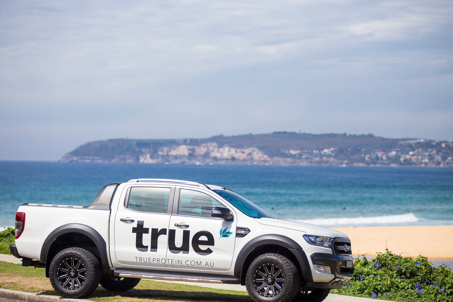 White Ford Ranger Wildtrak pickup truck with large "true" TrueProtein logo parked on grass beside an Australian coastal beach and ocean headland in the background.