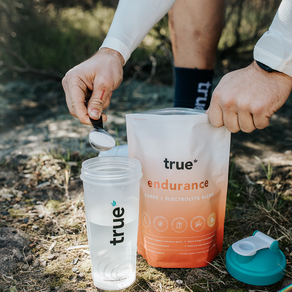 True Endurance carbs + electrolyte powder from True being scooped into a clear True shaker bottle outdoors on a trail, showing product pouch and shaker lid.