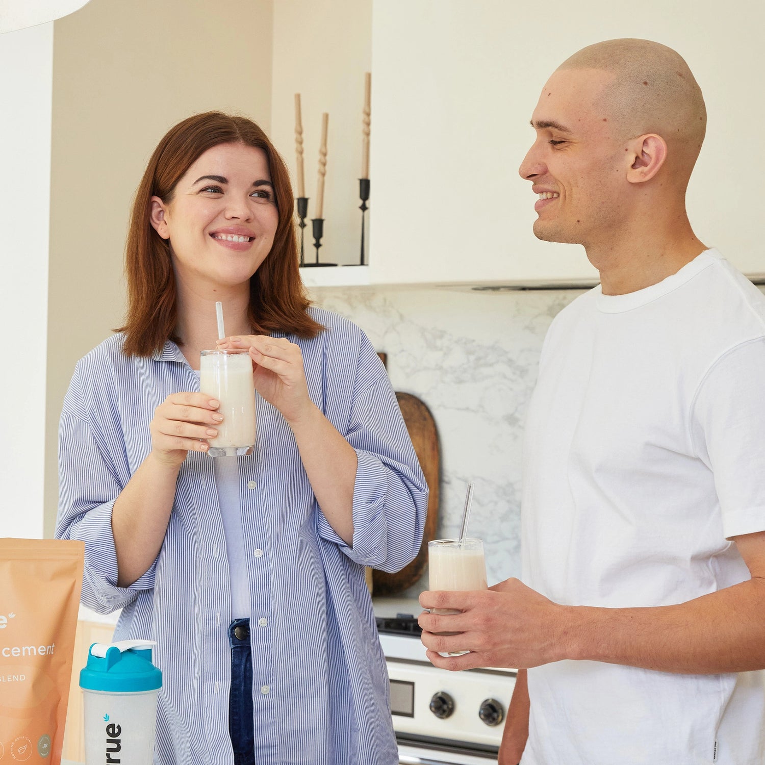 Two adults in a modern home kitchen smiling and holding creamy meal-replacement protein shakes with a True Protein bag and branded shaker visible on the counter — healthy protein shake breakfast lifestyle.