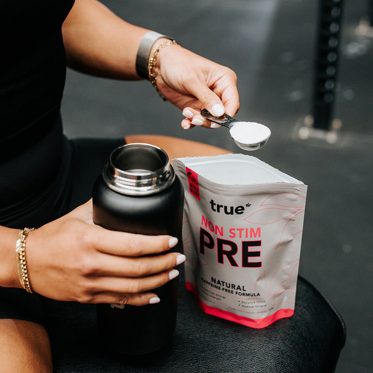 True Protein Non Stim PRE pre-workout powder being scooped into a black shaker bottle on a gym bench, natural caffeine-free formula.