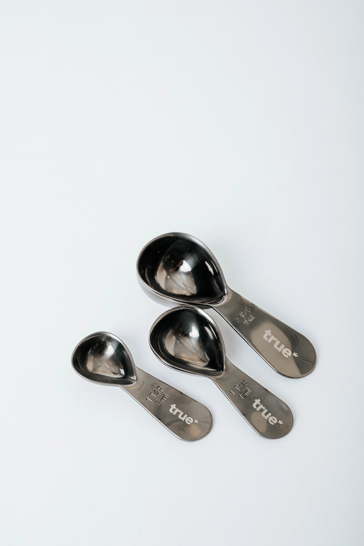 Three stainless steel measuring scoops (1 tsp/5 ml, 1 tbsp/15 ml, 2 tbsp/30 ml) with "true" logo on handles, arranged on a white background — sustainable protein scoop set product image.